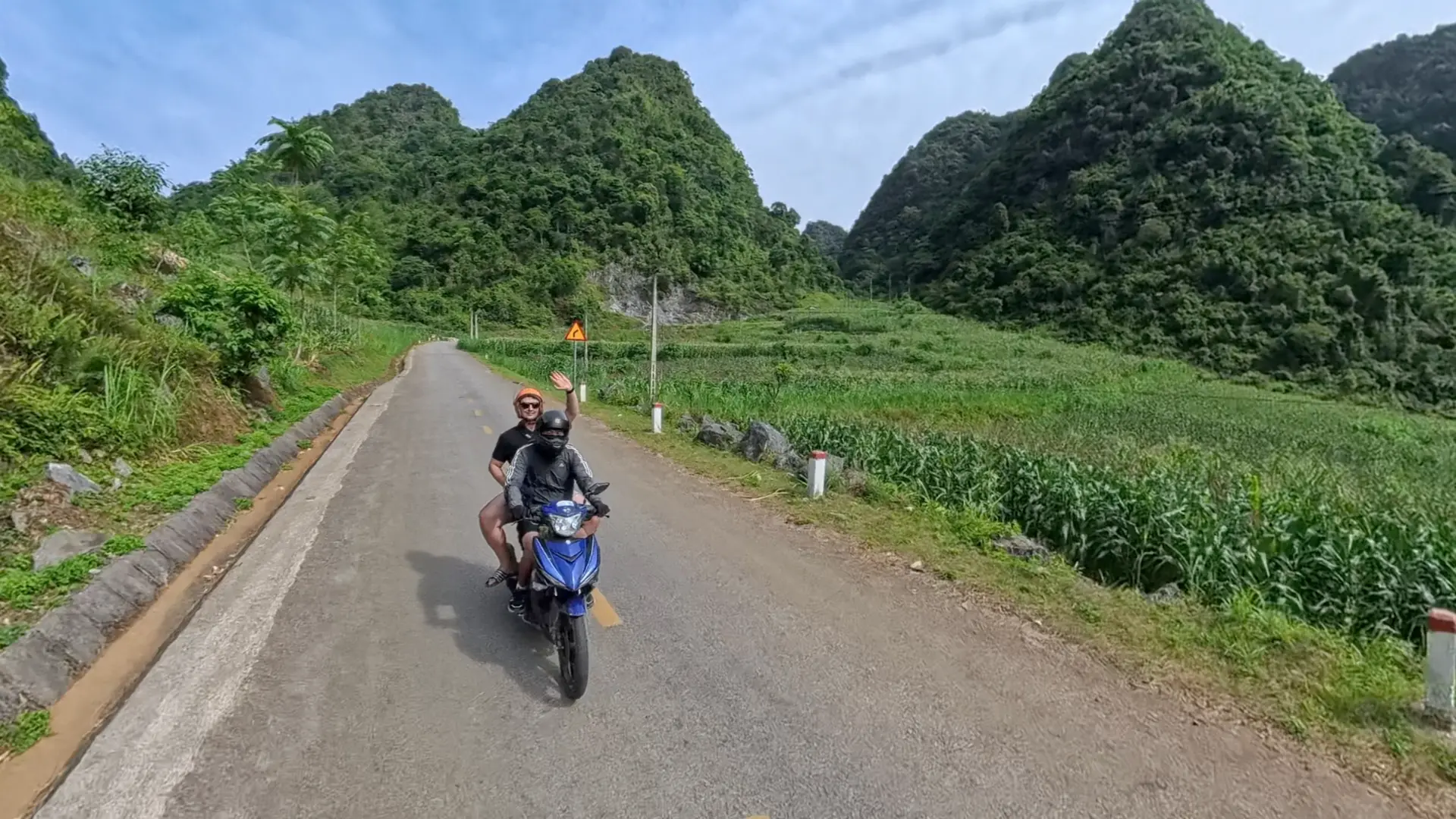 Traveler waving from the back of a blue motorbike on the Ha Giang Loop with green karst mountains behind, June 2026