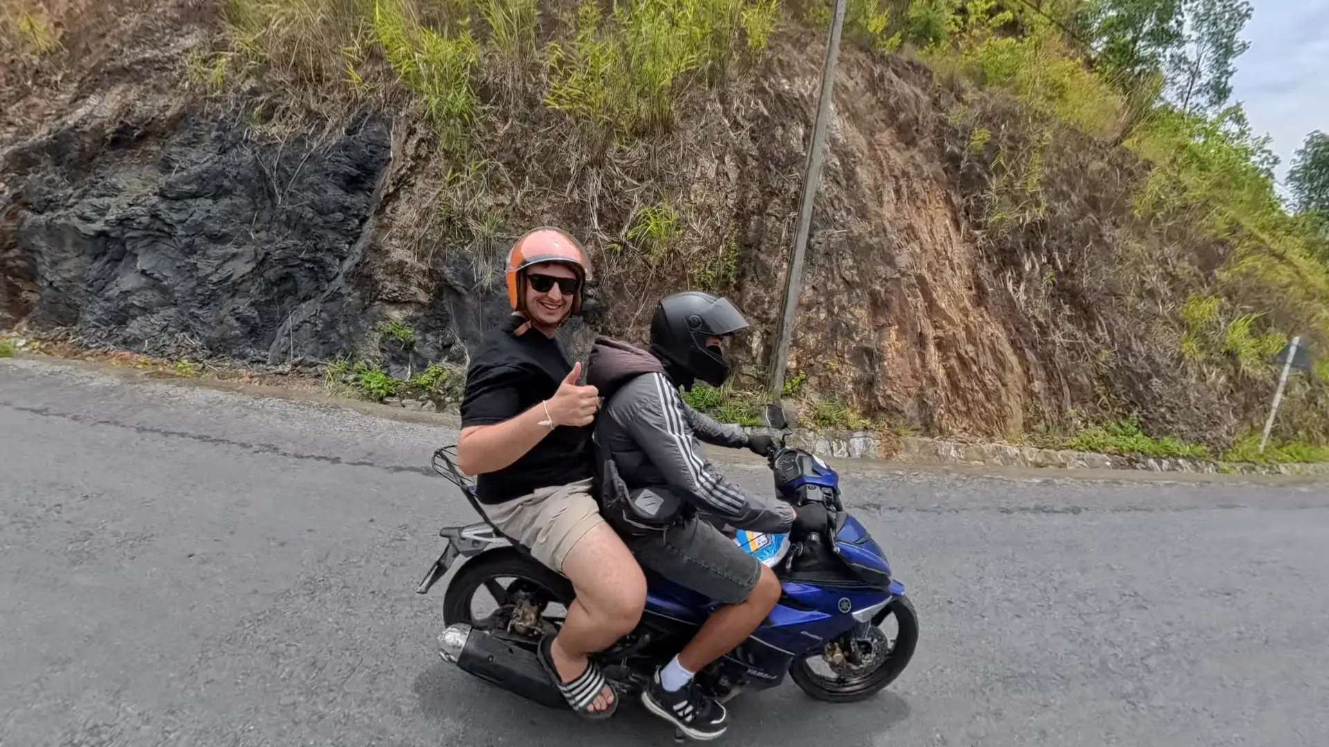 Traveler giving a thumbs up as passenger on a blue motorbike with an easy rider guide on a Ha Giang mountain road, June 2026