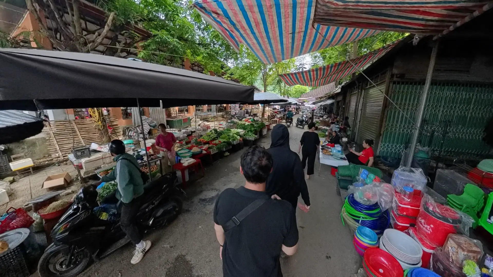 Walking through a busy local market in Ha Giang with produce stalls, plastic basins, and striped awnings, June 2026