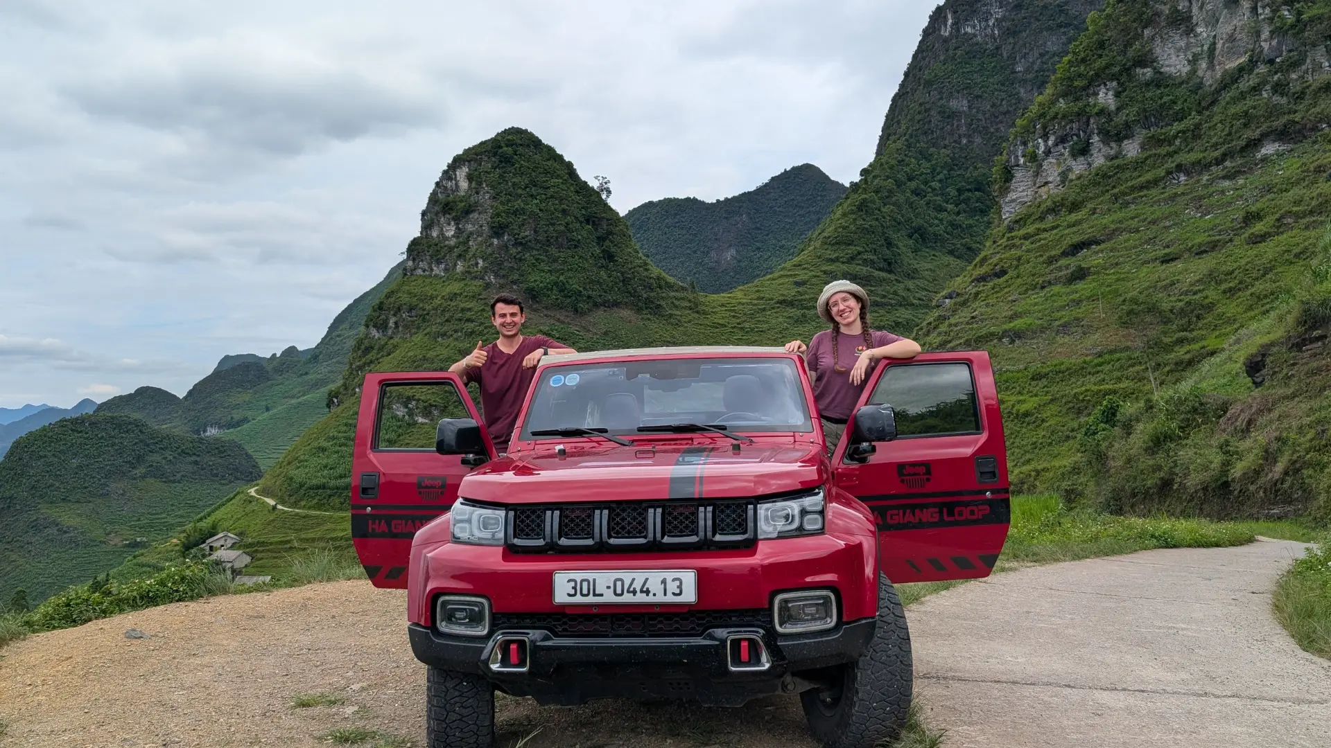 Liam and Sarah with a red Ha Giang Loop jeep on a mountain road overlooking green karst peaks, June 2026