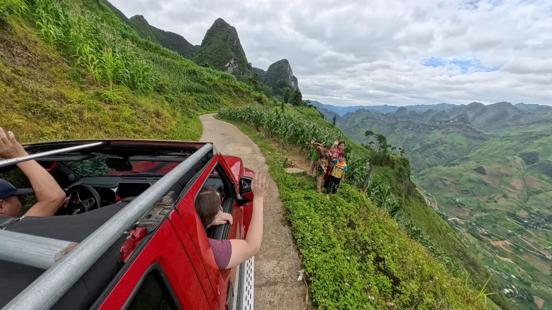 Passenger waving from a red open-air jeep to local children on a mountain road during the Ha Giang Loop, June 2026
