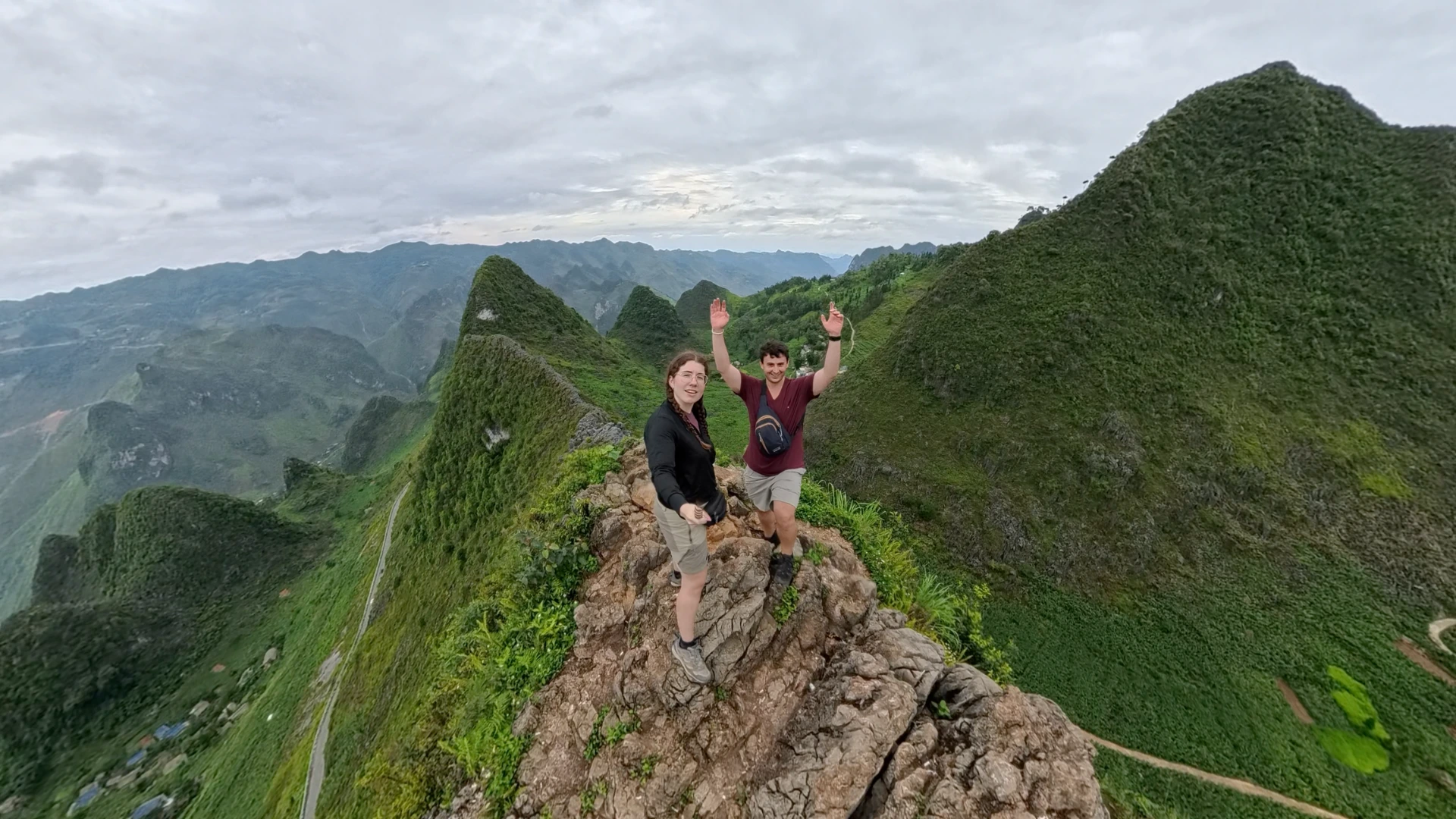 Liam and Sarah on a rocky ridge overlooking Ha Giang Loop karst mountains and a winding valley road, June 2026