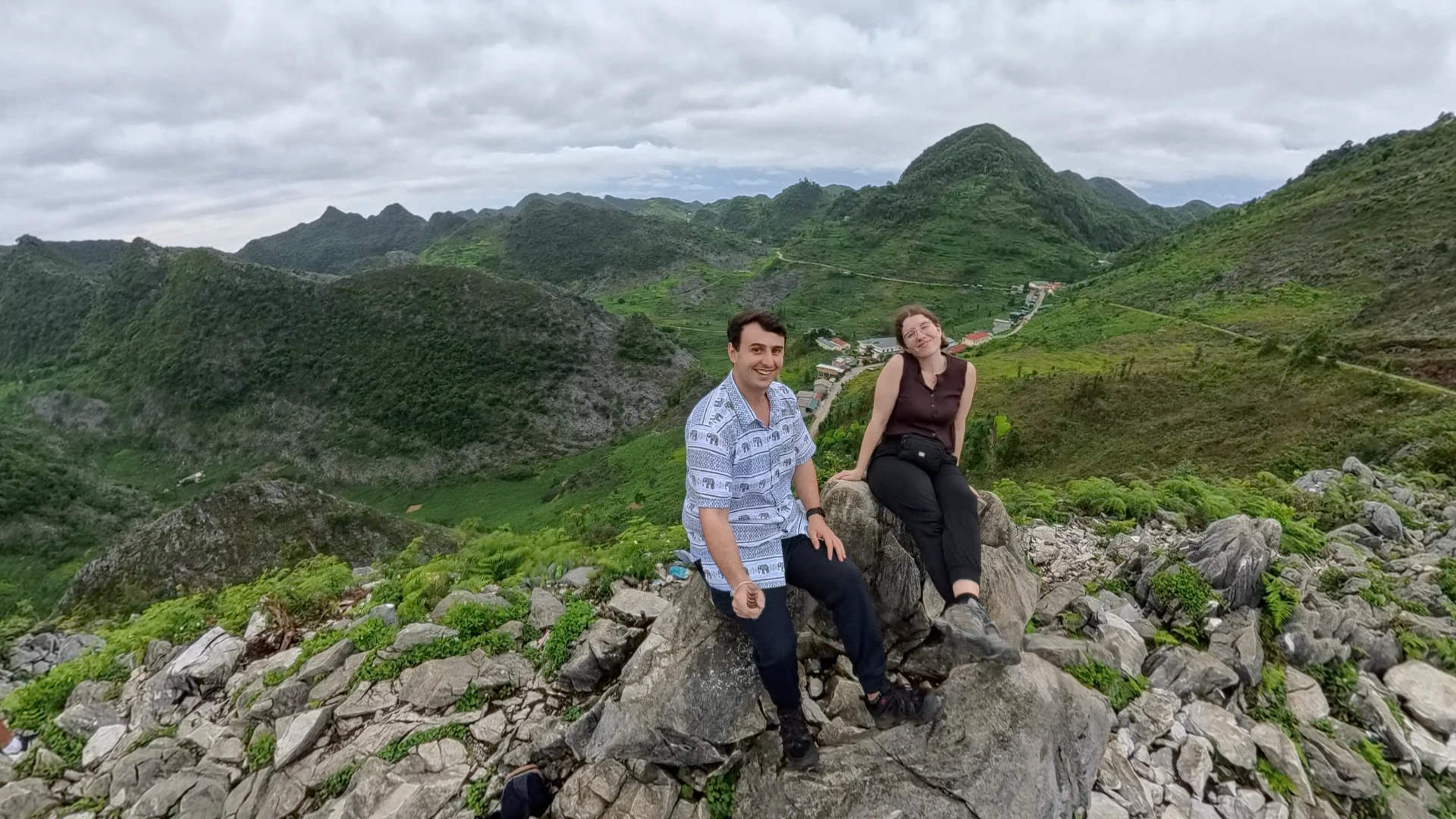 Liam and Sarah sitting on a rocky viewpoint overlooking green Ha Giang Loop mountains and a winding valley road, June 2026