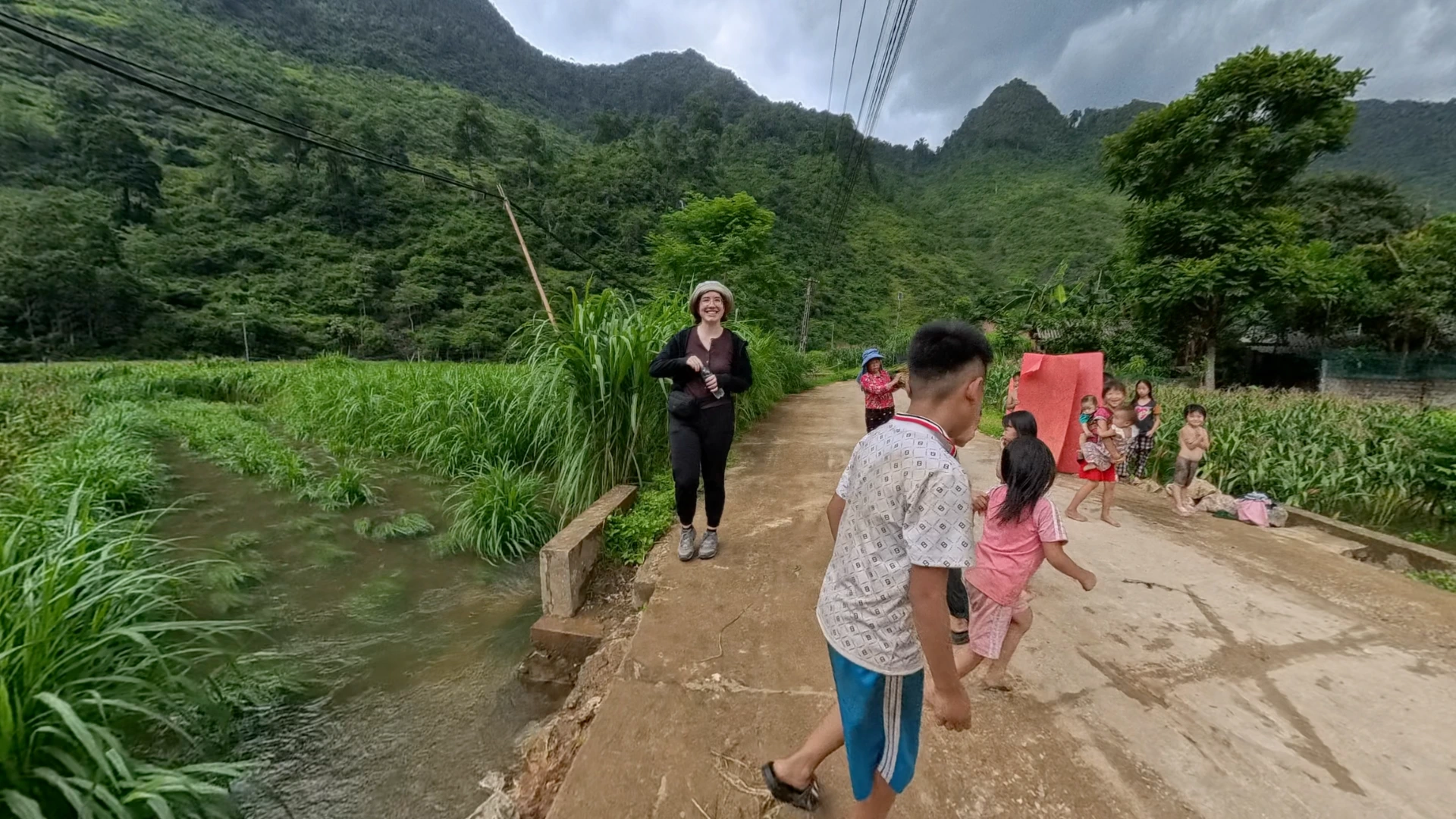 Sarah smiling on a rural Ha Giang road with local children playing nearby and karst mountains in the background, June 2026