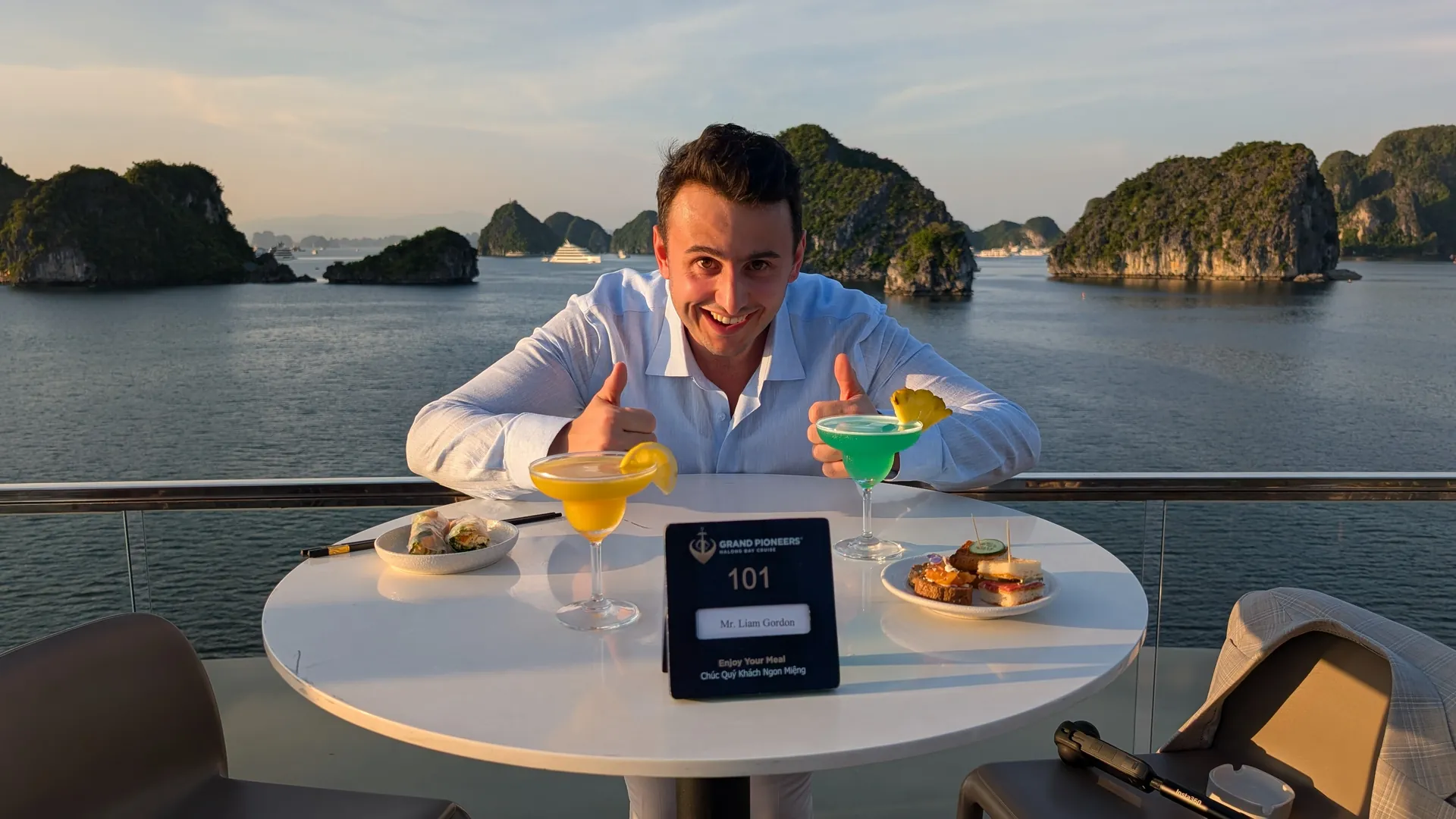 Liam giving two thumbs up at a sundeck table with colourful cocktails at sunset in Ha Long Bay