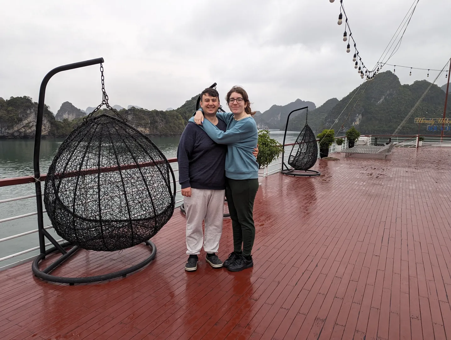 Liam and Sarah hugging on a rain-soaked cruise sundeck with egg chairs and misty Ha Long Bay cliffs