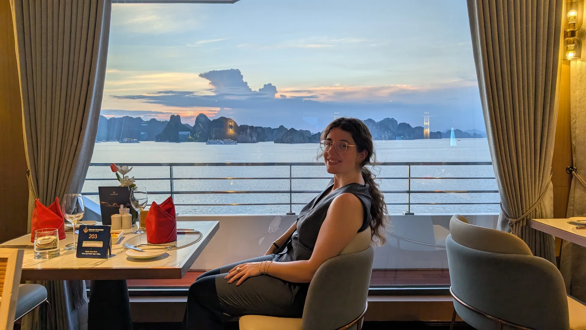 Sarah smiling at a dinner table by a big window as the sun sets over Ha Long Bay's limestone islands