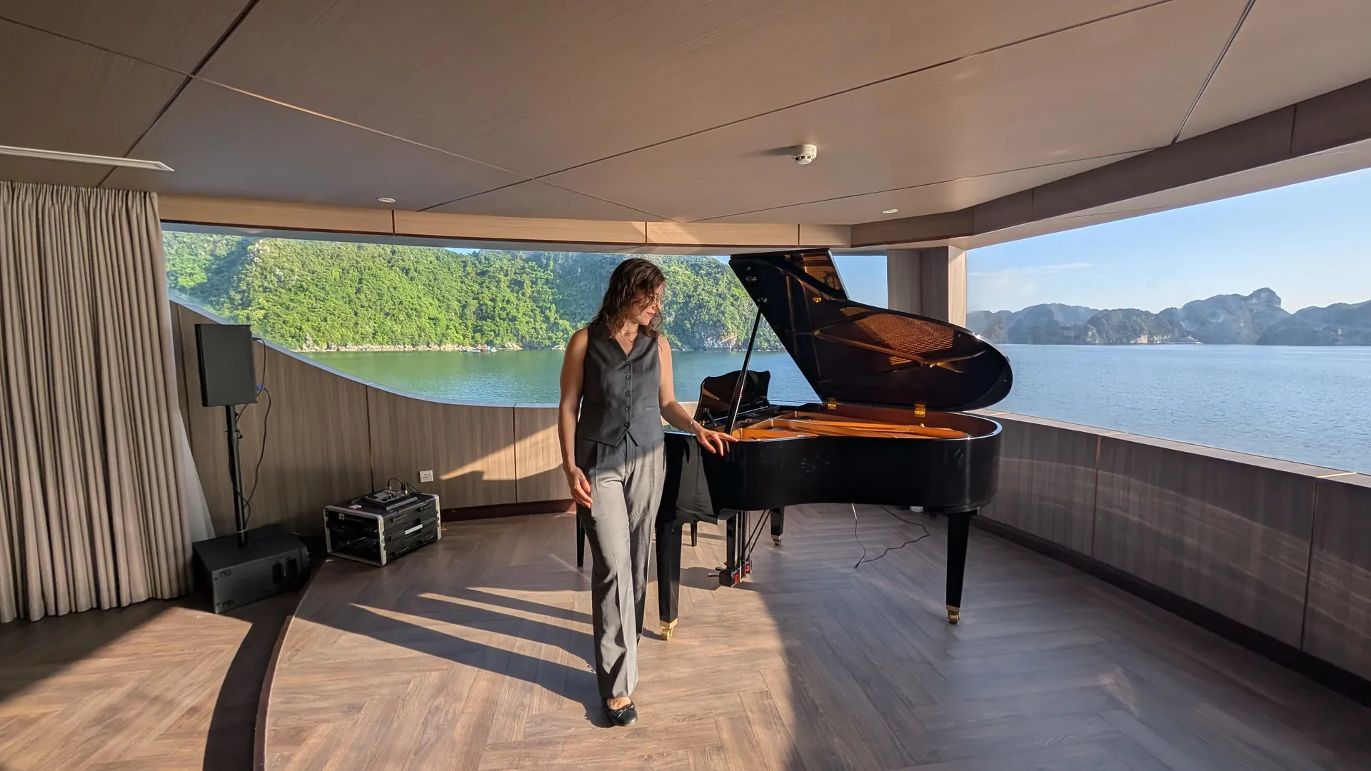 Sarah standing beside a grand piano in a cruise lounge with big windows looking out over Ha Long Bay