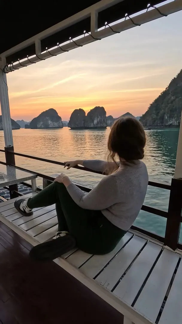 Sarah sitting on a wooden cruise deck watching the sun set behind Ha Long Bay's limestone islands