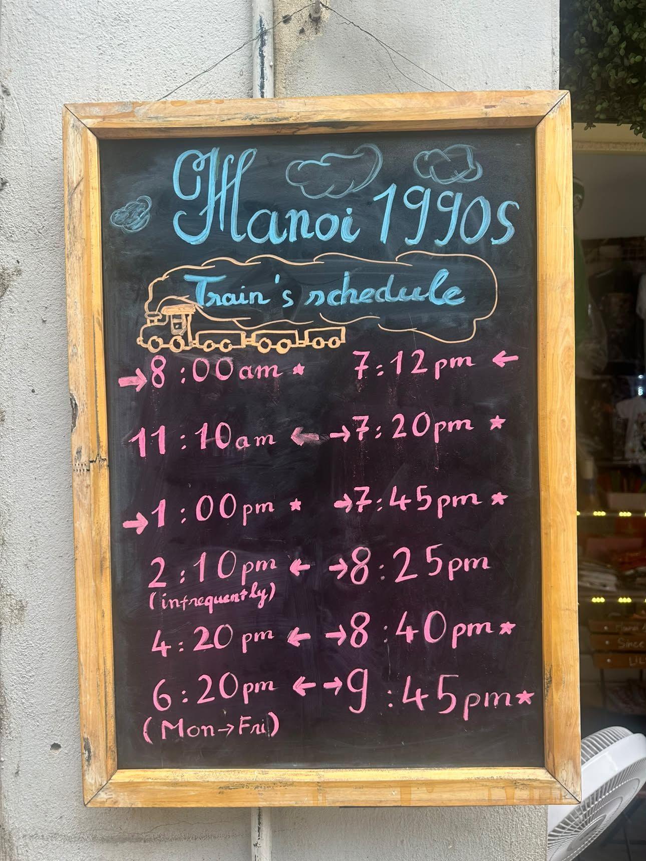Handwritten Train's schedule chalkboard at Hanoi 1990s cafe on Hanoi Train Street, wooden frame on a tiled wall, listing morning through evening pass times, photographed September 2026