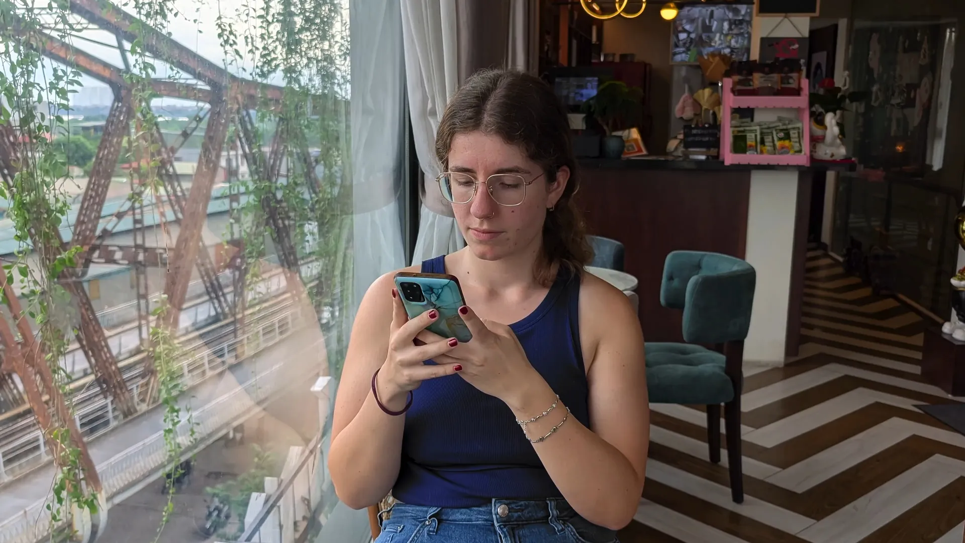 Sarah on her phone by a cafe window with a bridge outside, Vietnam, October 2026.
