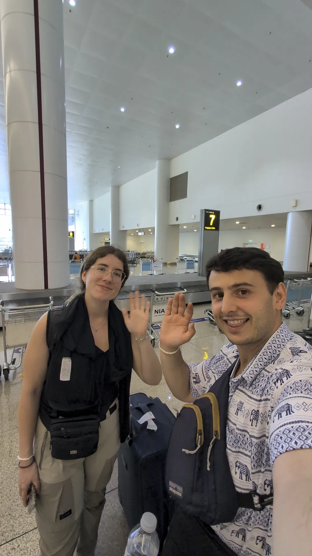Liam and Sarah smiling and waving next to their suitcase by the baggage belt at a Vietnam airport